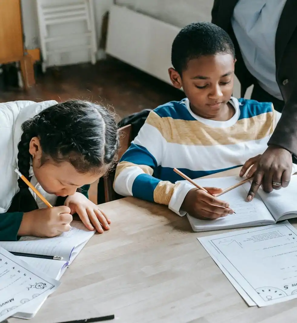 High angle of crop unrecognizable black female teacher explaining task to focused little schoolboy sitting at desk near attentive Asian classmate writing in notebook